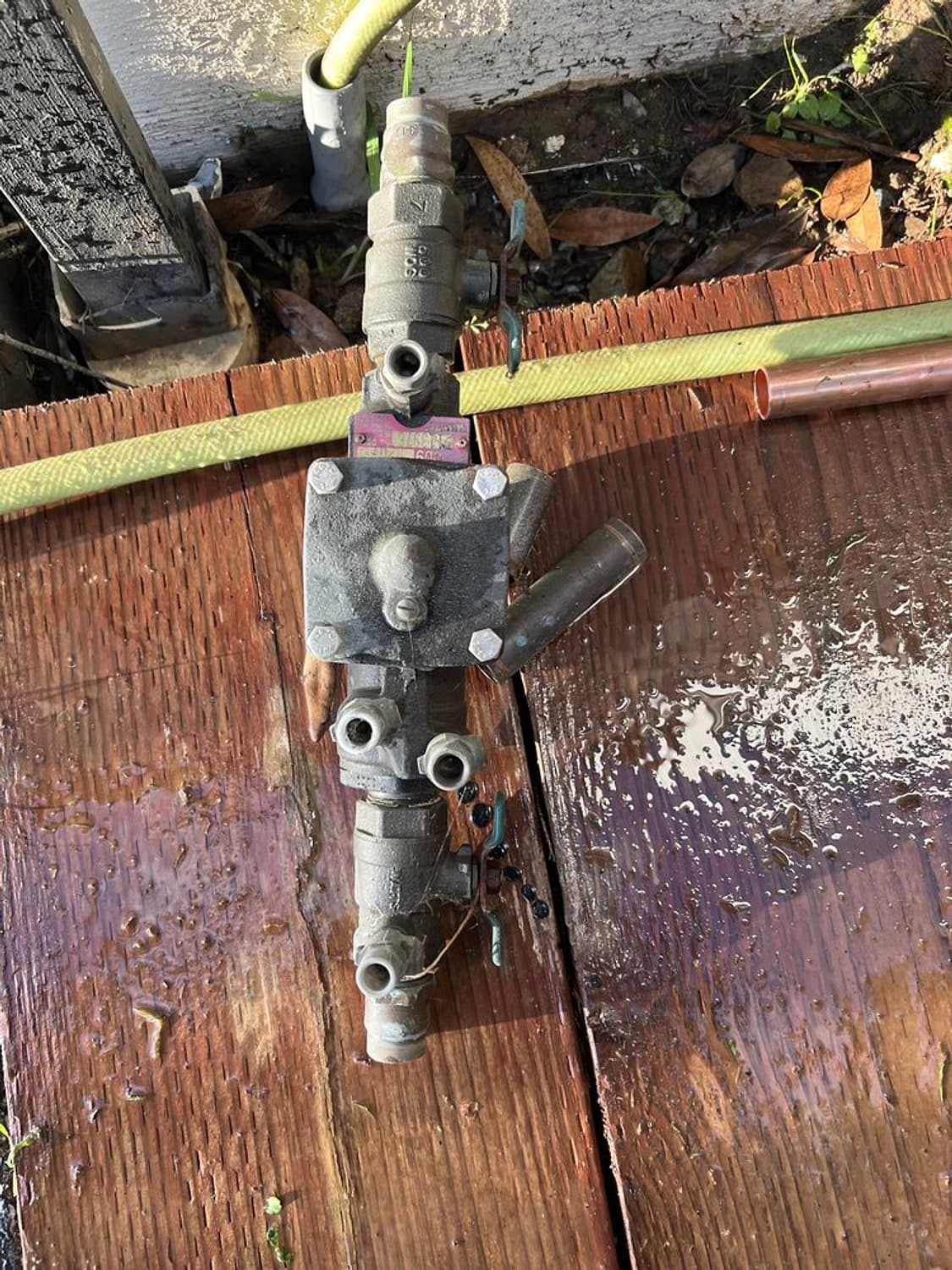 Old rusted backflow valve assembly taken out and lying on a wet wooden deck beside a garden hose