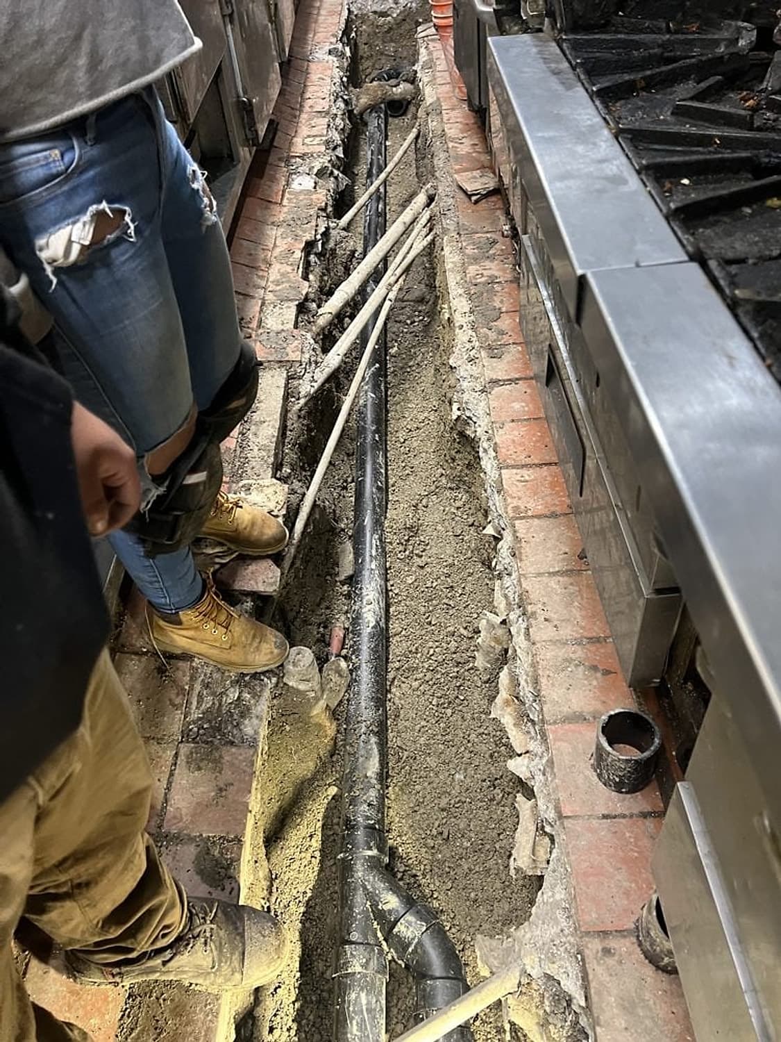 Zanja abierta en el piso de una cocina comercial con una línea de drenaje nueva de ABS negro junto al equipo de cocina