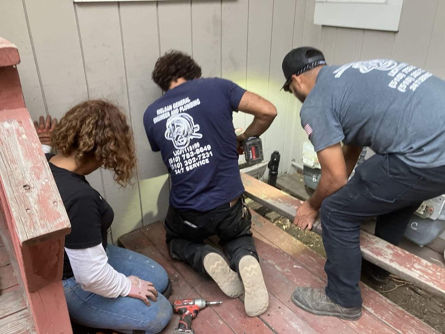 Three people kneeling on a floor during a repair, one in an AB’s Plumbing shirt, with tools and an open floor section