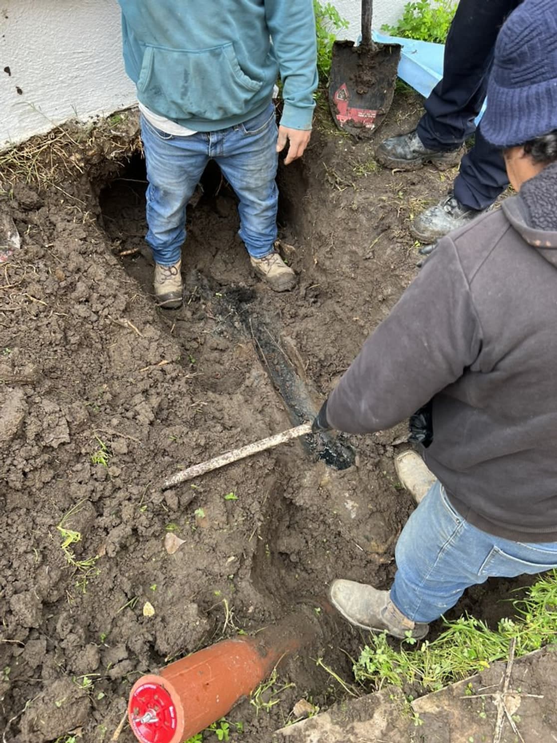 Two crew members in a trench beside a house, pulling out an old clay sewer line, with a piece of clay pipe in the foreground