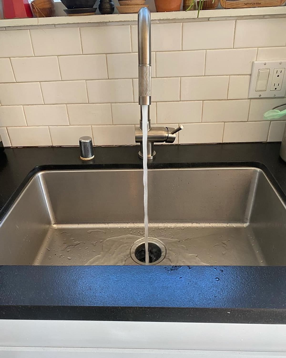 New tall stainless kitchen faucet running into a deep stainless sink, with white tile behind