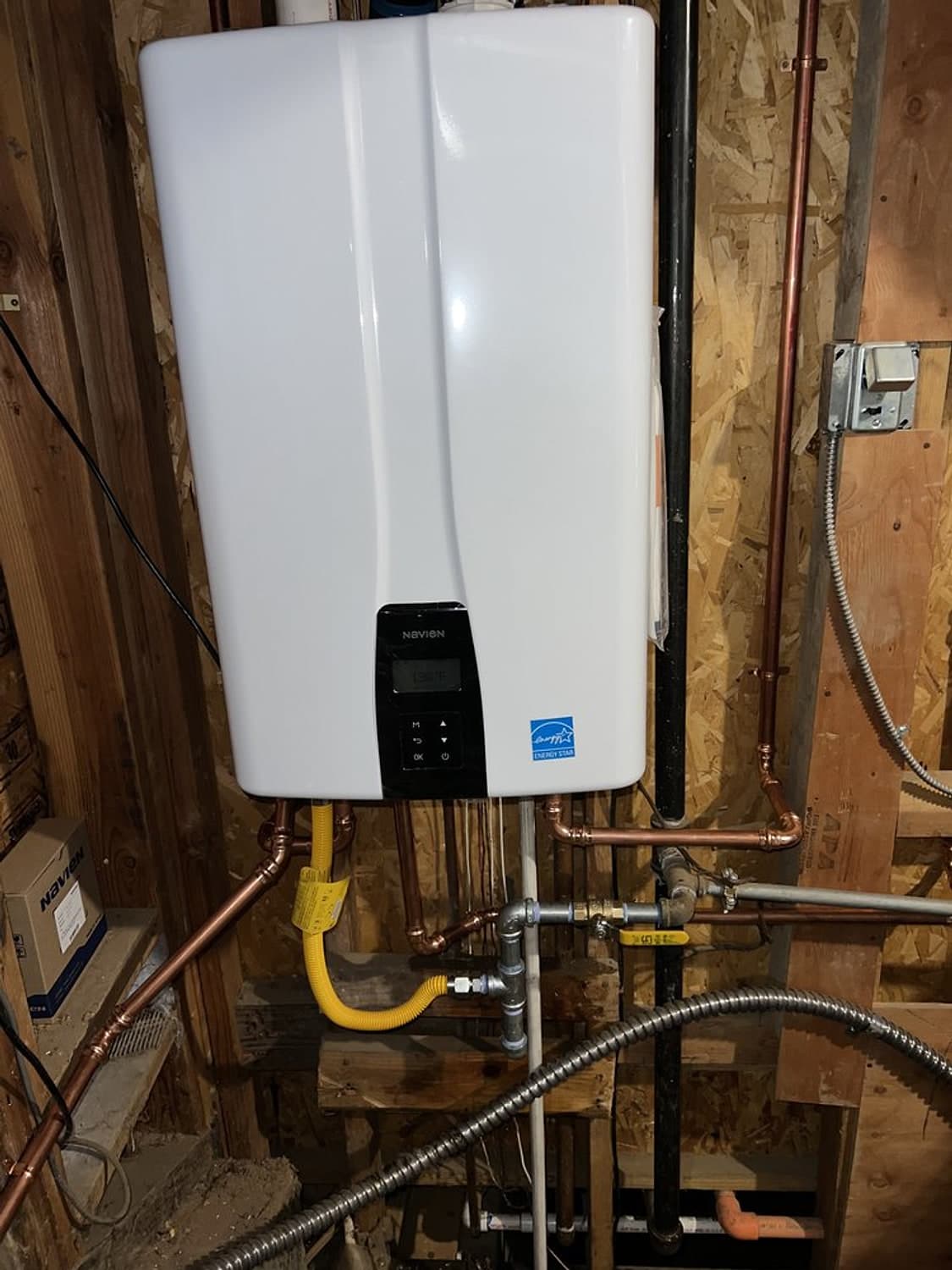 White Navien tankless water heater on a wooden wall, with a yellow gas line and new water connections below