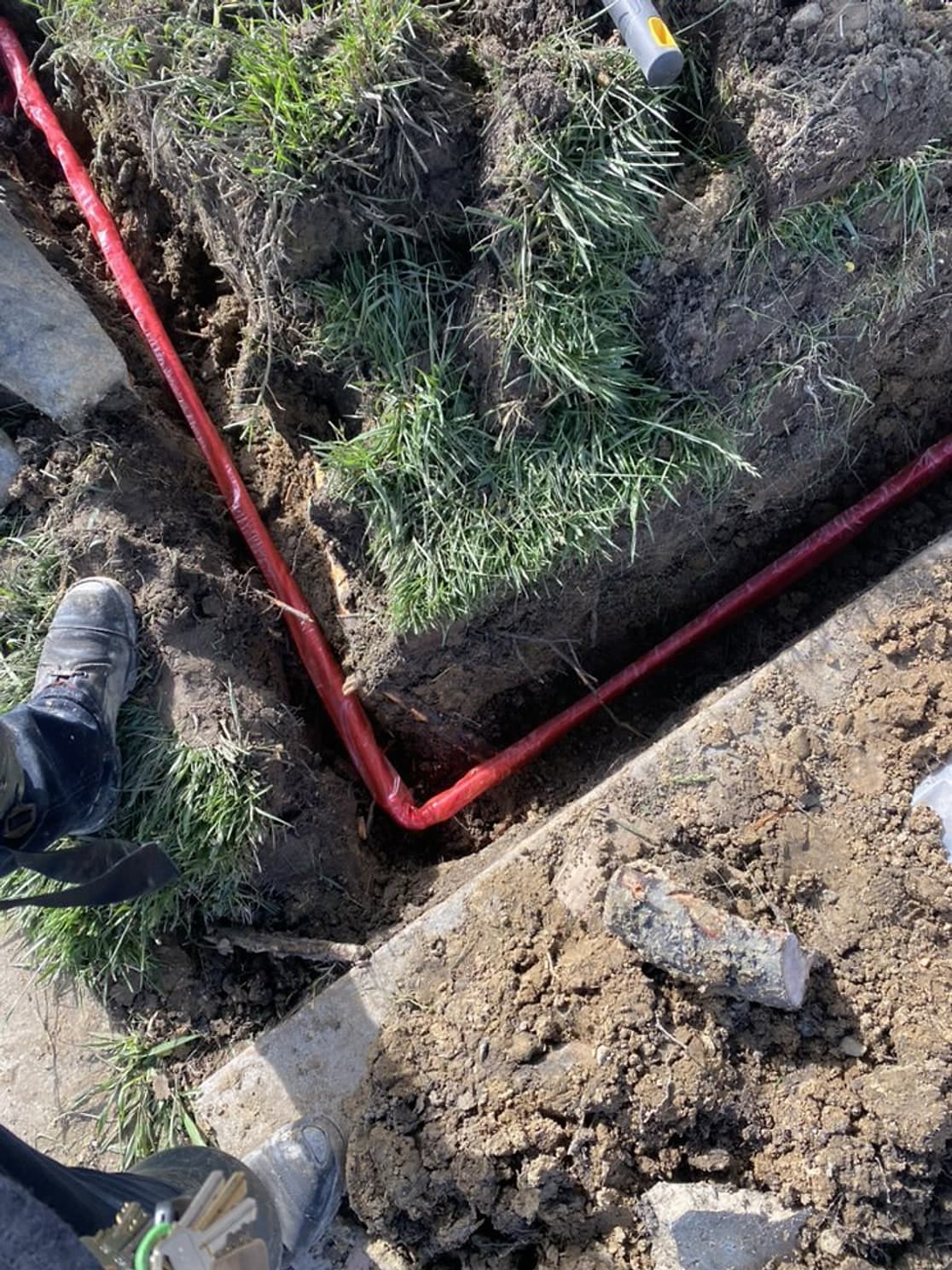 A new red-sleeved main water line in a trench turning a corner along a walkway