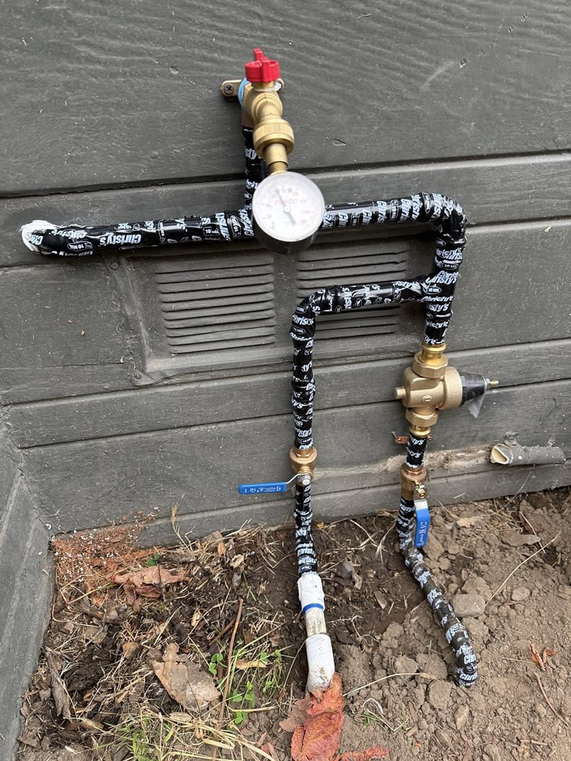 Brass pressure regulator, pressure gauge and red shutoff valve on a wrapped water line against a gray house wall
