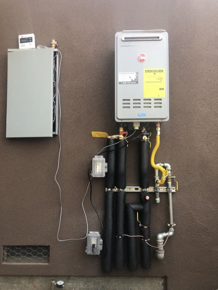 Rheem tankless water heater on a brown exterior wall with black insulated pipes and a yellow gas line