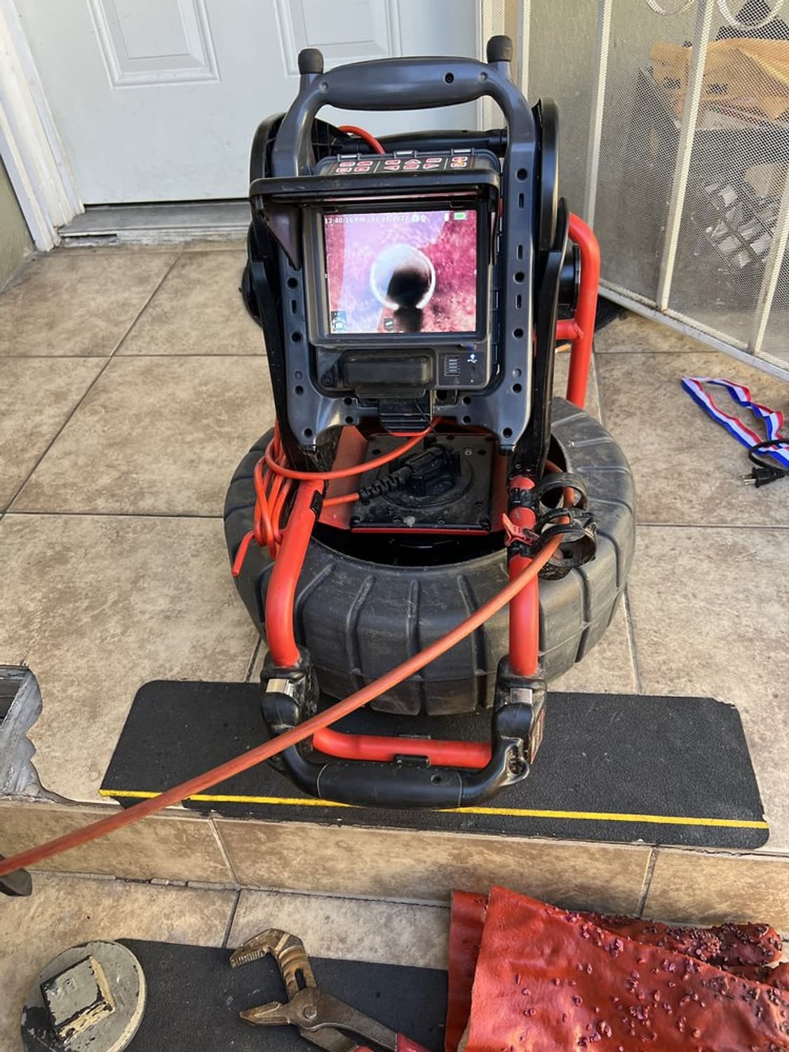 Sewer inspection camera reel and monitor set up on a floor mat, with the cable running to the drain