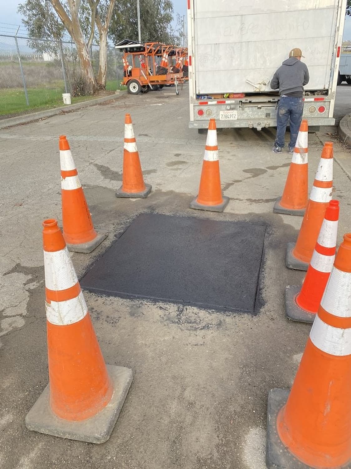 Parche de asfalto nuevo en la calle rodeado de conos naranjas después de conectar una línea lateral de drenaje, con un camión de trabajo atrás