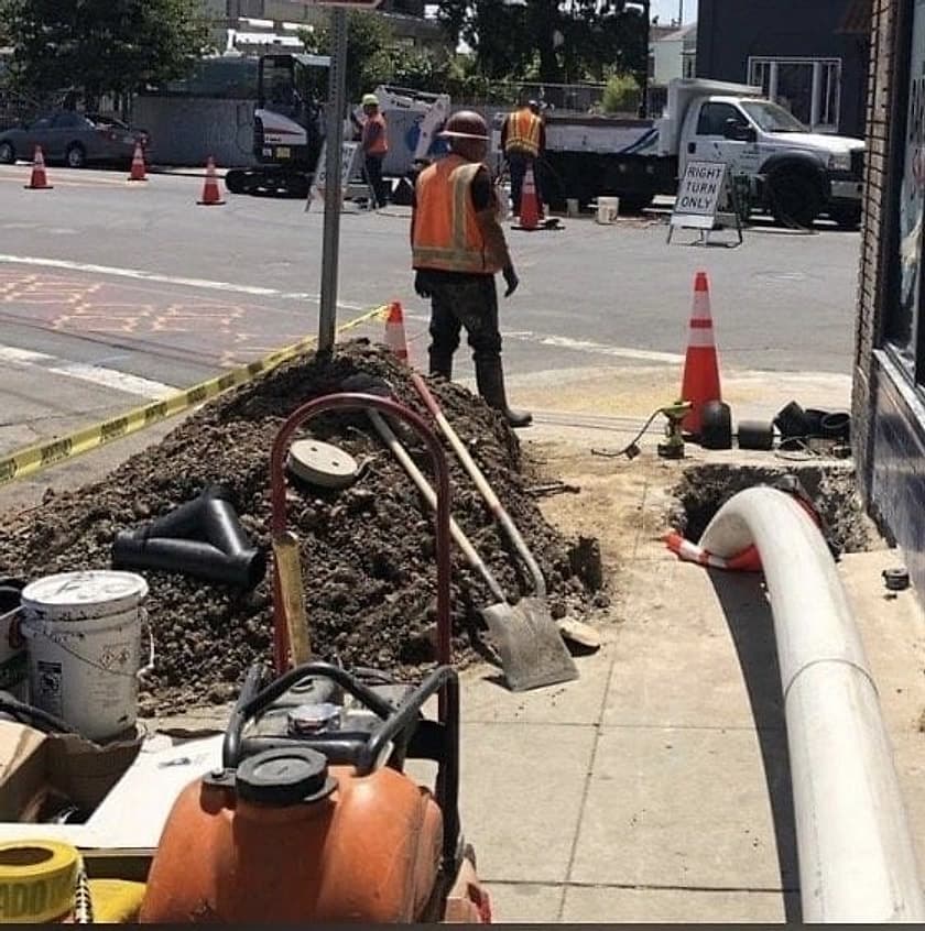 Crew in hard hats and hi-vis vests at an open trench with new white pipe on Foothill Blvd in Oakland, with traffic cones and a work truck behind