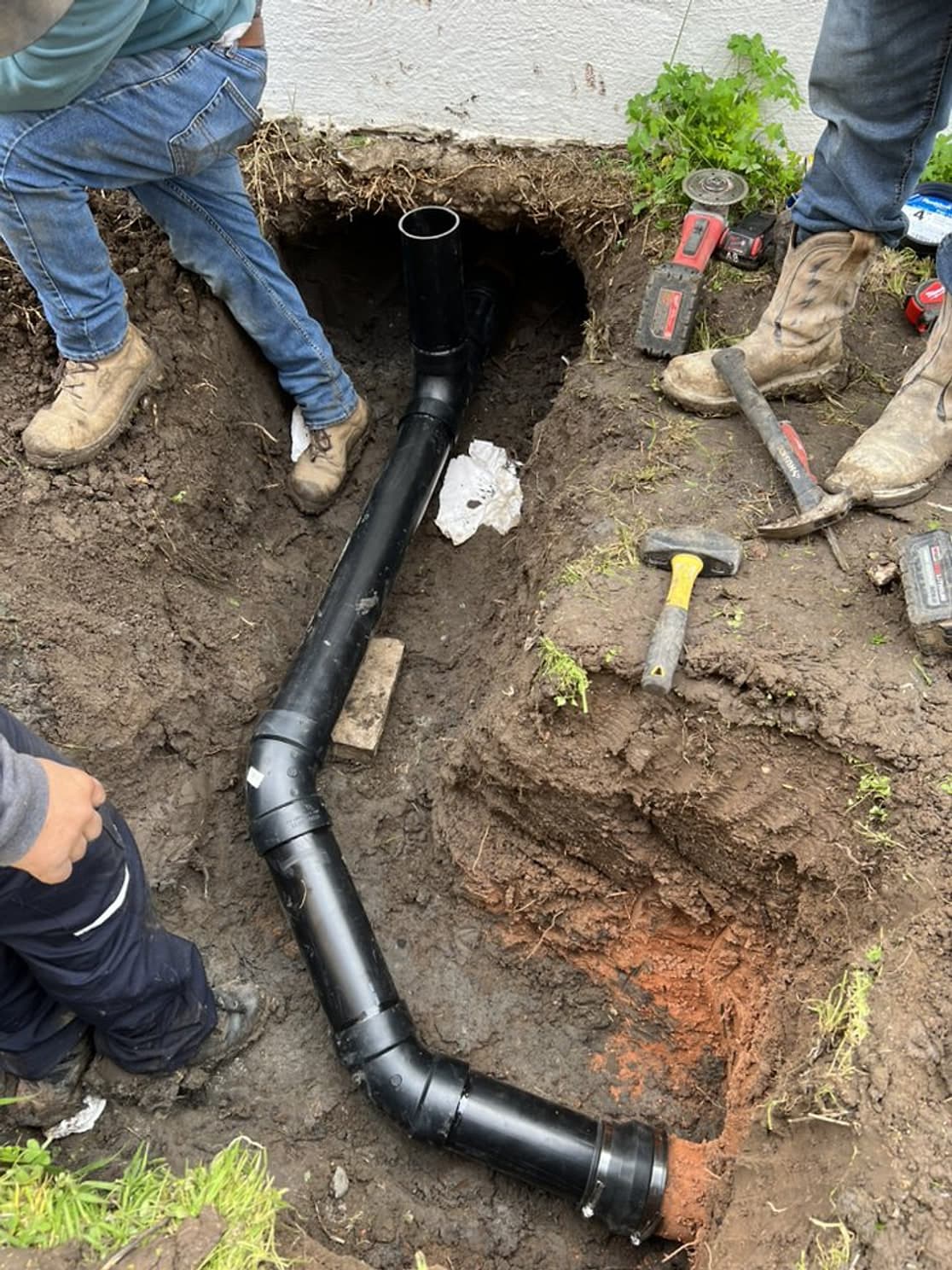 Crew members around an open trench where a new black ABS sewer line runs from the house, with shovels and tools nearby