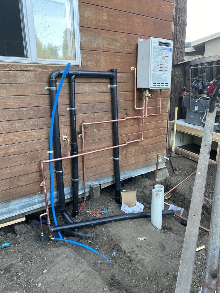 Tankless water heater on wood siding with new black drain pipes, copper and blue water lines running into the ground