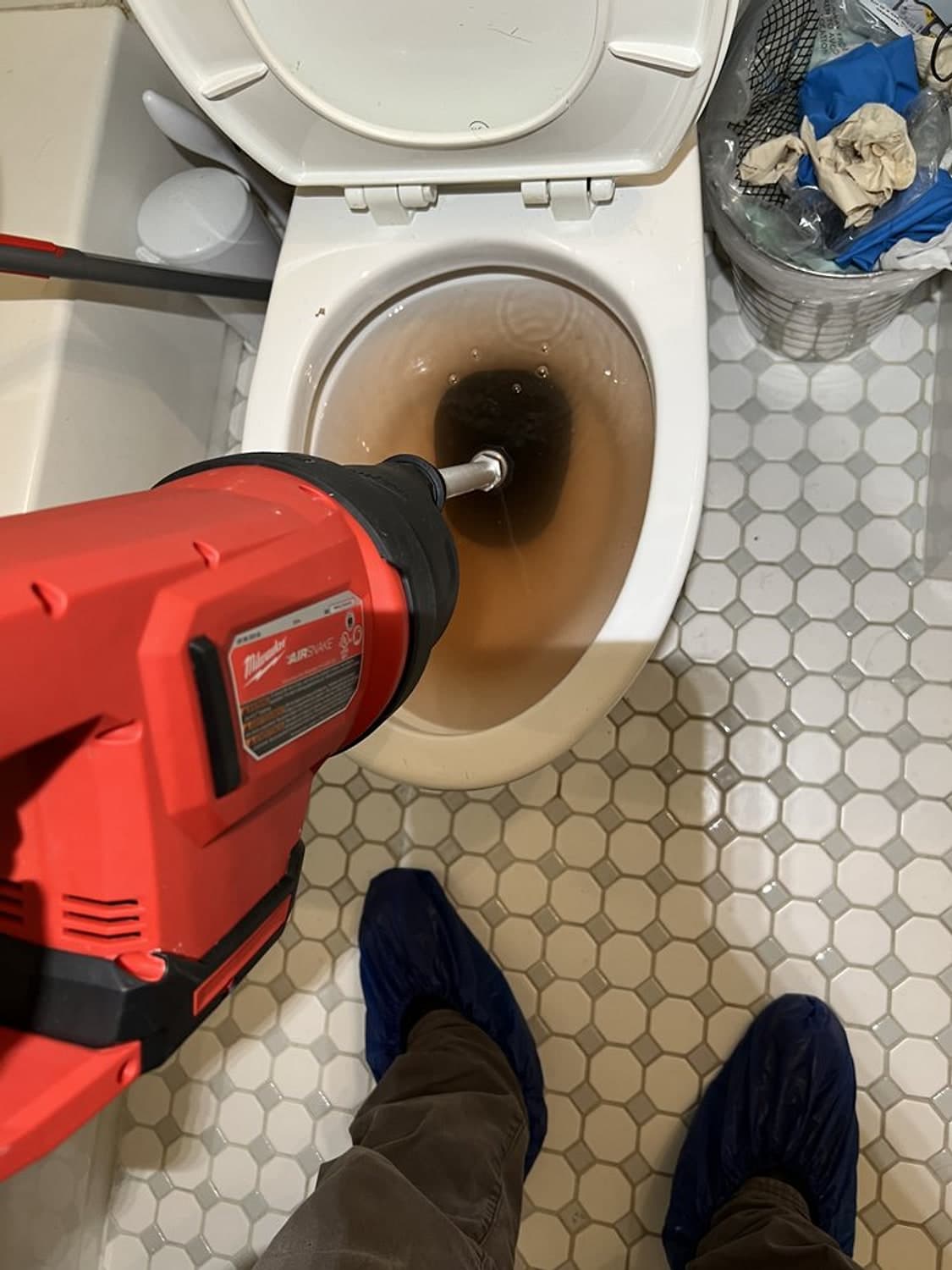 A red power drain auger fed into a clogged toilet, plumber wearing shoe covers