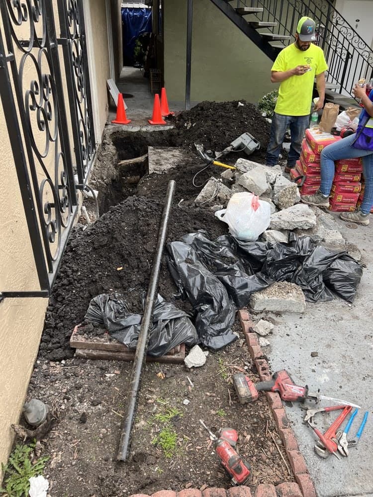 Long trench dug beside an apartment building with an old pipe exposed, tools and traffic cones nearby