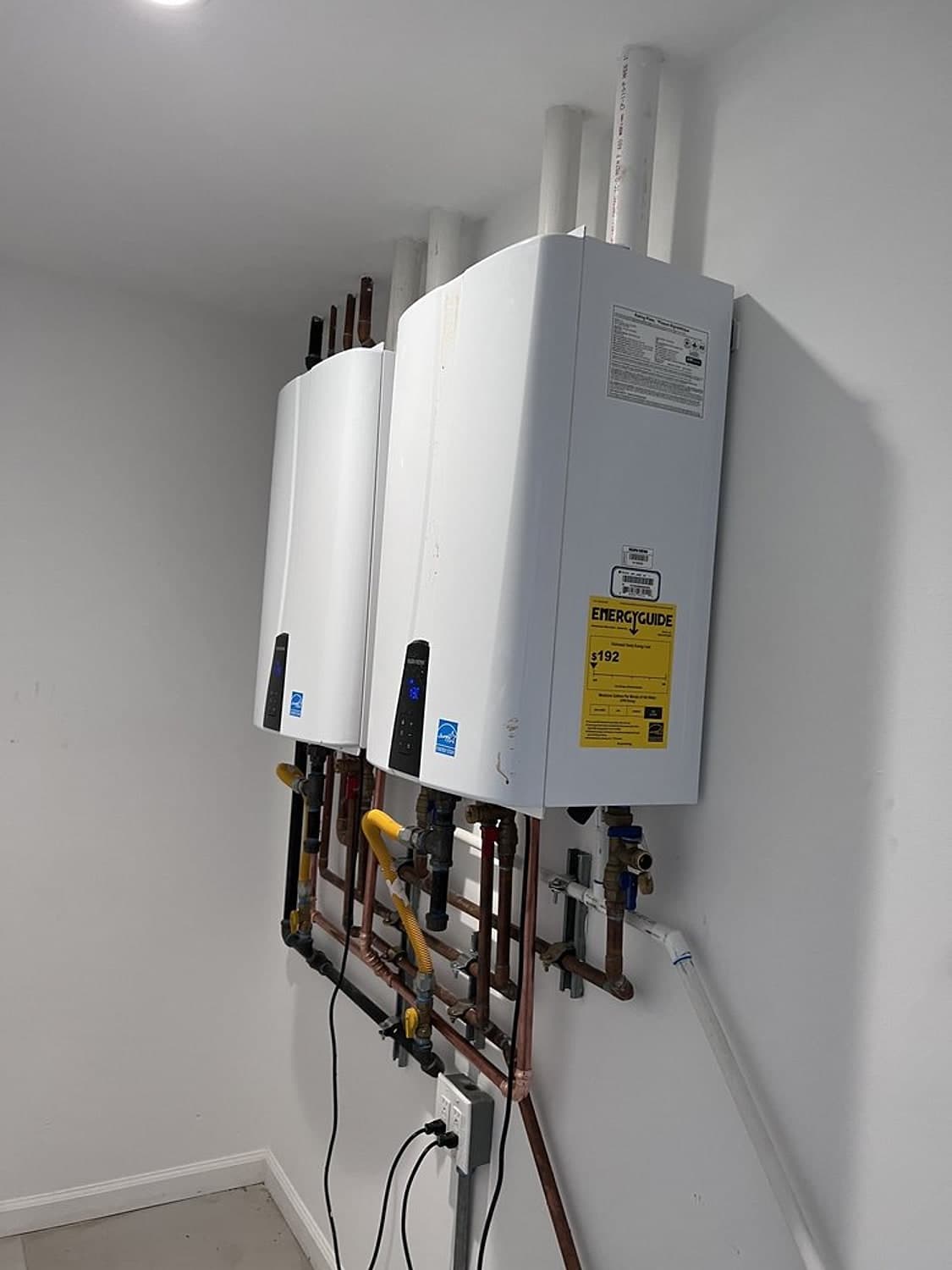 Two white tankless water heaters mounted side by side on a wall, with new vent pipes and copper lines below