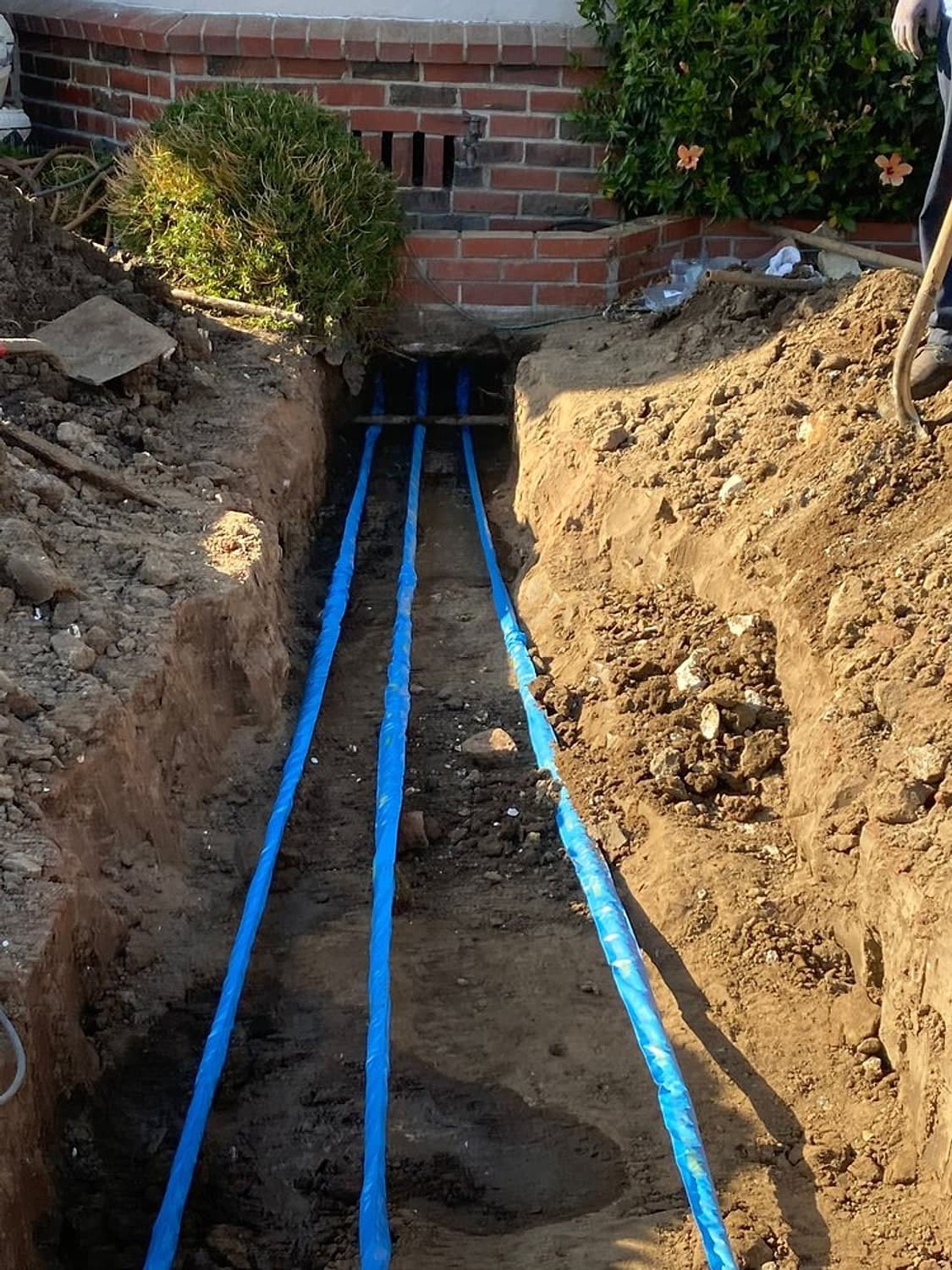 Two new blue-sleeved water lines laid in a long open trench beside a brick wall
