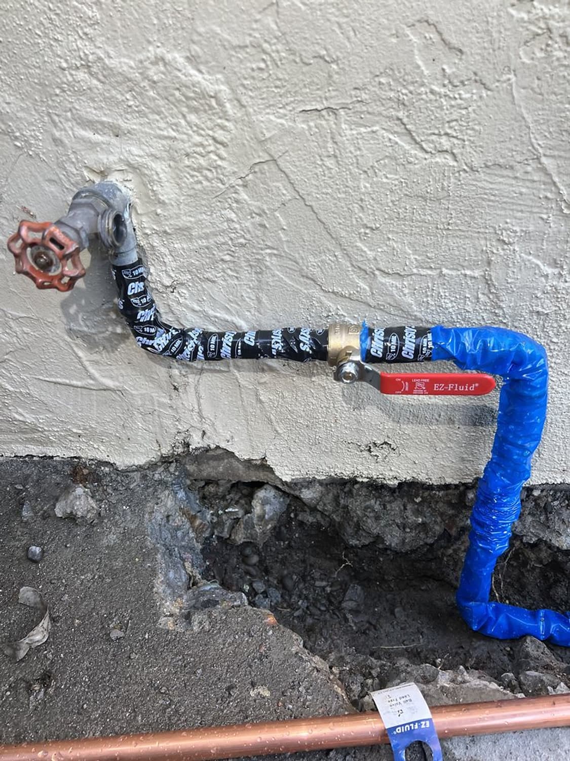 New water line wrapped in blue protective tape with a red shutoff valve, entering a stucco wall above an open trench